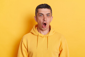 Wall Mural - Portrait of surprised man with dark hair wearing casual style hoodie standing isolated over yellow background, sees something astonishing, keeps mouth widely opened.