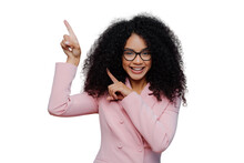 Half Length Shot Of Cheerful Afro American Woman With Dark Bushy Hairstyle, Wears Violet Suit, Points Above With Both Index Fingers, Demonstrates Way To Something, Invites You Going Upstairs