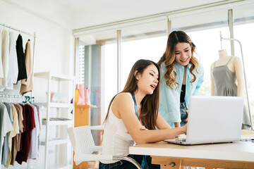 Two Asian female fashion designers or dressmakers brainstormed and discuss the design of the new collection with examples of fabrics and sketches on the laptop. Working together in the studio.