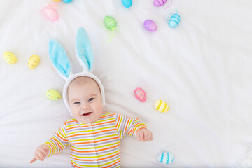 Wall Mural - happy baby boy with rabbit ears on his head lying on the bed with Easter eggs, cute funny smiling little baby. The concept of Easter. Space for text