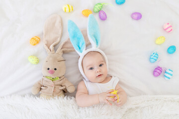 Wall Mural - A baby boy with rabbit ears on his head is lying in a crib with a bunny toy and Easter eggs, a cute funny smiling little baby. Easter Concept