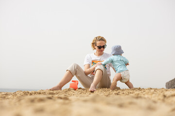 Wall Mural - Mother playing his infant baby boy son on sandy beach enjoying summer vacationson on Lanzarote island, Spain. Family travel and vacations concept