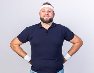 Wall Mural - smiling adult slavic sporty man wearing headband and wristbands putting hands on waist and looking at camera isolated on white background with copy space