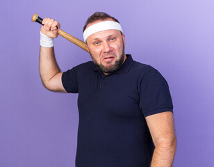 Wall Mural - annoyed adult slavic sporty man wearing headband and wristbands holding bat on shoulder isolated on purple background with copy space