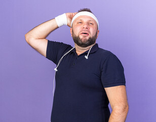 Wall Mural - pleased adult slavic sporty man with headphones around neck wearing headband and wristbands putting hand on head and looking at side isolated on purple background with copy space