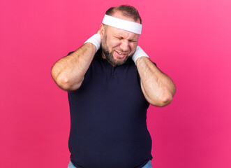 Wall Mural - aching adult slavic sporty man wearing headband and wristbands holding his neck with hands isolated on pink background with copy space