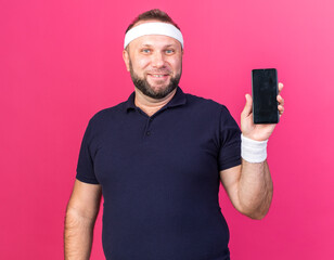 Wall Mural - smiling adult slavic sporty man wearing headband and wristbands holding phone isolated on pink background with copy space