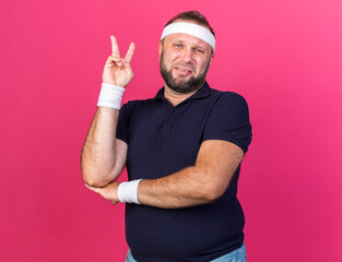 Wall Mural - aching adult slavic sporty man wearing headband and wristbands gesturing victory sign isolated on pink background with copy space