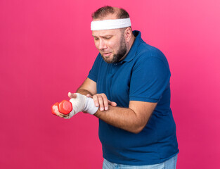 Wall Mural - sad adult slavic sporty man wearing headband and wristbands holding dumbbell and looking at his hand isolated on pink background with copy space