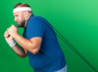 Wall Mural - unpleased adult slavic sporty man wearing headband and wristbands pulls jumping rope isolated on green background with copy space