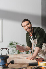 Wall Mural - Smiling carpenter in apron holding sandpaper near board and looking at camera in workshop.