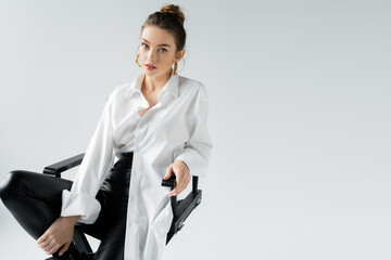 trendy woman in black tight pants and white oversize shirt sitting on chair and looking at camera isolated on grey.