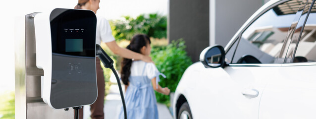 Focus electric charging station power by clean energy for electric vehicle at home with blurred progressive woman and girl walking in background. Home charging station for electric engine car concept