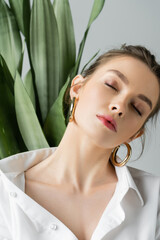 Wall Mural - portrait of sensual woman in white shirt and hoop earrings posing with closed eyes near blurred plant isolated on grey.