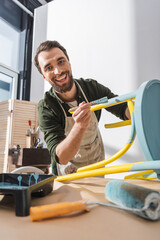 Wall Mural - Smiling restorer looking at camera while holding paintbrush near chair in workshop.