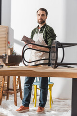 Wall Mural - Restorer holding sandpaper and looking at camera near wooden chair in workshop.