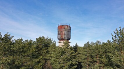 A huge water tank, a water tower taller than a pine forest, stands in a dense forest. Rusty abandoned soviet water tower in Russia against the blue sky.