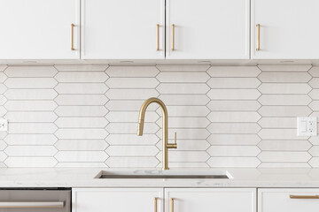 A beautiful kitchen faucet detail with white cabinets, a gold faucet, white marble countertops, and a brown picket ceramic tile backsplash.