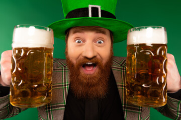 Wall Mural - Portrait of excited man in hat holding beer glasses isolated on green.