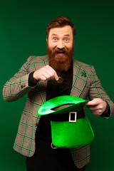 Wall Mural - Smiling bearded man looking at camera while holding coin and hat isolated on green.