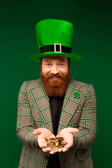 Wall Mural - Smiling bearded man in hat holding coins while celebrating saint patrick day isolated on green.