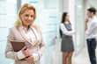 © Bojan - Portrait of beautiful middle aged blond women in elegant suit with notebook standing in office hall.