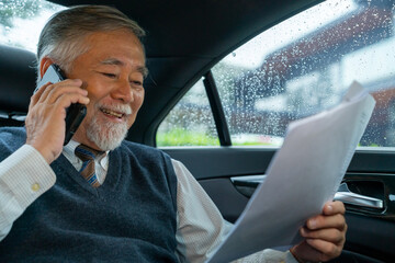 Confident senior business man in suit sitting on car backseat with reading business plan document and talking on mobile phone during go to office. Elderly businessman executive working on the road.