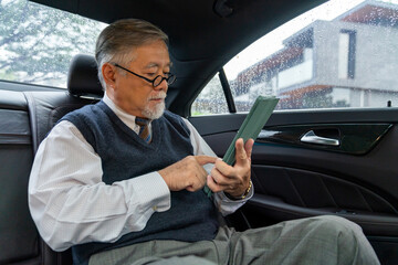 Confident senior business man in suit sitting on car backseat with working on digital tablet and drinking hot coffee during go to office. Elderly businessman executive working on the road.