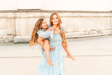 Wall Mural - Mother of the daughter walks playing. Mother holds the girl on her back, holding her legs, and her daughter hugs her by the shoulders. Dressed in blue dresses.