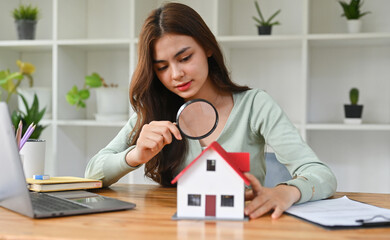 Beautiful young woman holding magnifying glass and looking at house model. Real estate appraisal, ouse search concept.