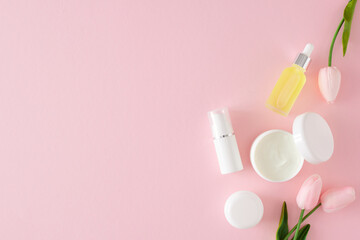 Wall Mural - Skincare beauty concept. Flat lay photo of white cosmetic tubes, cream jars, serum bottles and spring flowers on pastel pink background with copyspace. Skin care products mockup.