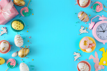 Wall Mural - Easter card concept. Flat lay photo of color eggs and easter candy with chocolate sprinkles cute rabbits with gift box pink alarm clock on pastel blue background with blank space in the middle.