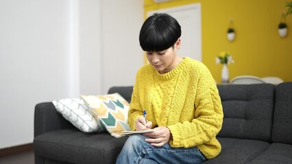 Wall Mural - Young chinese woman writing on notebook sitting on sofa at home