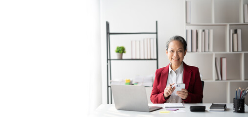 Wall Mural - Senior Asian businesswoman in bright red suit using her smartphone while working in the modern office room.