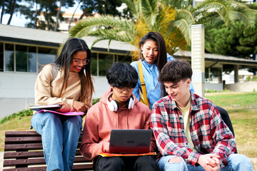 Young international university students happily stare at the computer. Classmates looking at the laptop screen. Teenagers reviewing for exam or working on group project sitting on a high school bench.