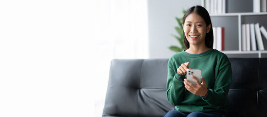 Wall Mural - Adorable young Asian girl using her smartphone, surfing the internet while relaxing on her sofa at home.