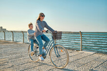 Happy Family, Carefree Mother And Son With Bike Riding On Beach Having Fun, On The Seaside Promenade On A Summer Day, Enjoying Vacation. Togetherness Friendly Concept
