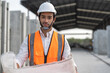 © Supachai - Confident man engineer wearing protective helmet and vest holding blueprint and smiling at factory or construction site making precast concrete wall for real estate housing.