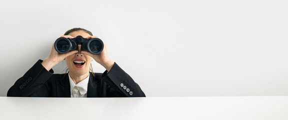 Wall Mural - Surprised young woman looking through binoculars peeking out from behind white cardboard. Banner