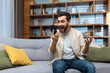 © Liubomir - Mature man celebrating victory and successful achievement, got news online message on app using phone sitting on sofa in living room.