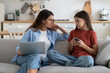 © DimaBerlin - Teen girl daughter hiding messages on phone from curious mother sitting beside on sofa, selective focus. Family mom and child resting at home with gadgets. Parental control on smartphone