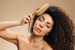 © SHOTPRIME STUDIO - HEALTHY LONG HAIR CARE CONCEPT. Happy cheerful beautiful tanned awesome curly Latin lady with hairbrush combing hair posing isolated on pastel beige background, closing eyes. Copy space, cool offer