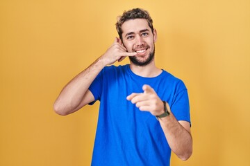 Wall Mural - Hispanic man with beard standing over yellow background smiling doing talking on the telephone gesture and pointing to you. call me.