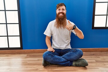 Canvas Print - Redhead man with long beard sitting on the floor at empty room doing happy thumbs up gesture with hand. approving expression looking at the camera showing success.