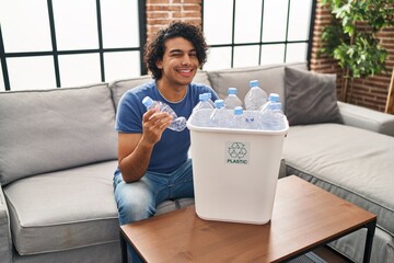 Wall Mural - Hispanic man with curly hair holding recycling bin with plastic bottles at home winking looking at the camera with sexy expression, cheerful and happy face.