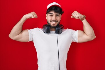 Canvas Print - Hispanic man with beard wearing gamer hat and headphones showing arms muscles smiling proud. fitness concept.