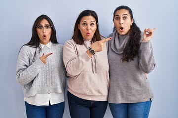 Canvas Print - Mother and two daughters standing over blue background surprised pointing with finger to the side, open mouth amazed expression.