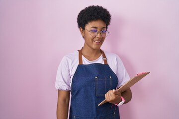 Sticker - Young african american woman wearing professional waitress apron holding clipboard winking looking at the camera with sexy expression, cheerful and happy face.