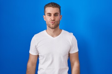 Wall Mural - Young caucasian man standing over blue background looking at the camera blowing a kiss on air being lovely and sexy. love expression.