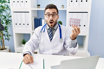 Sticker - Handsome hispanic man wearing doctor uniform holding prescription pills scared and amazed with open mouth for surprise, disbelief face